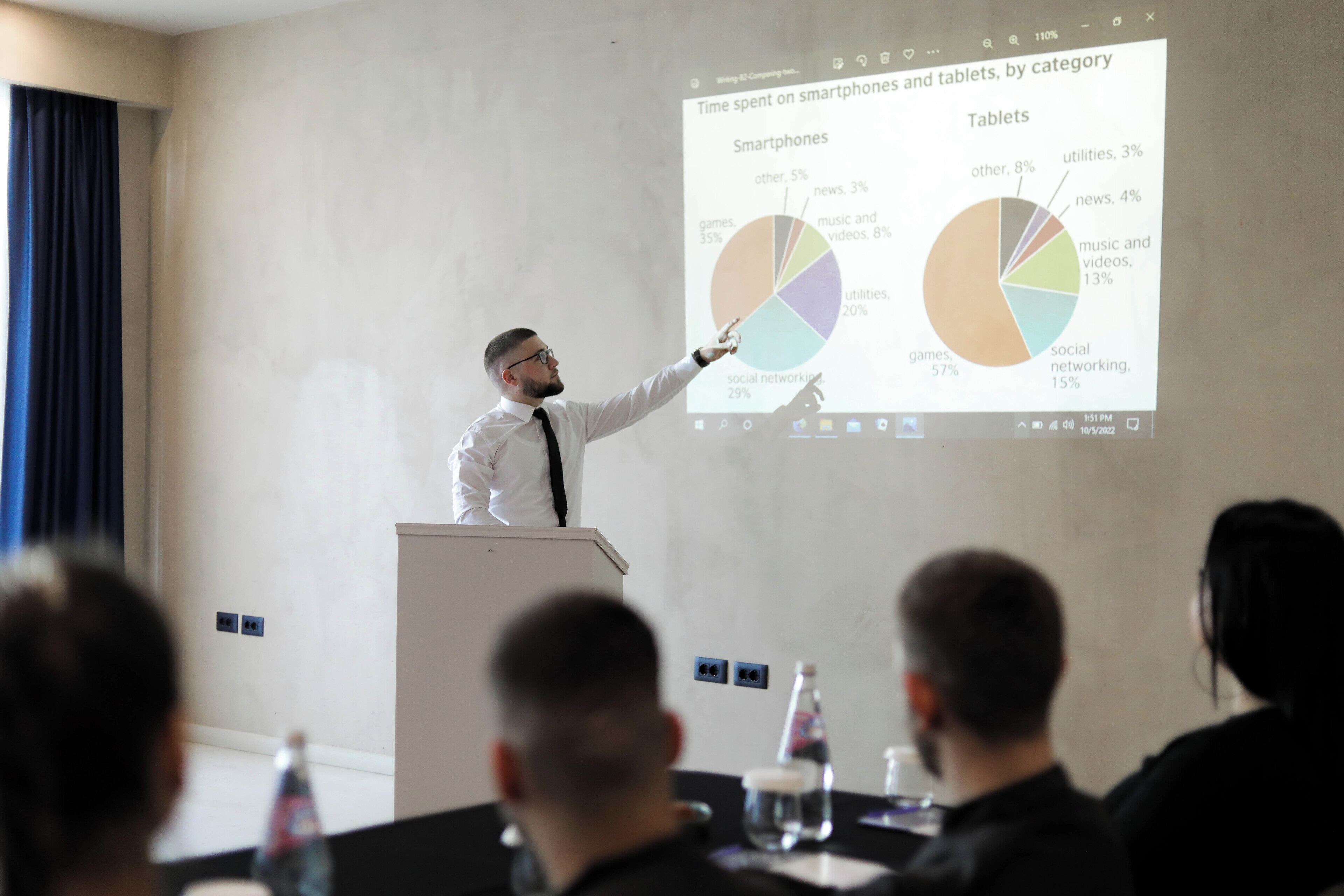 A presentation with a speaker at a podium, a pie chart on the screen, and an audience seated at a table with water bottles.