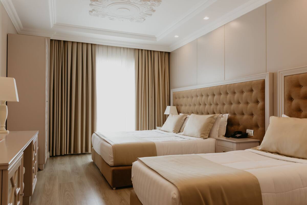 A hotel room with two beds, a nightstand, a dresser, and a large window with curtains.