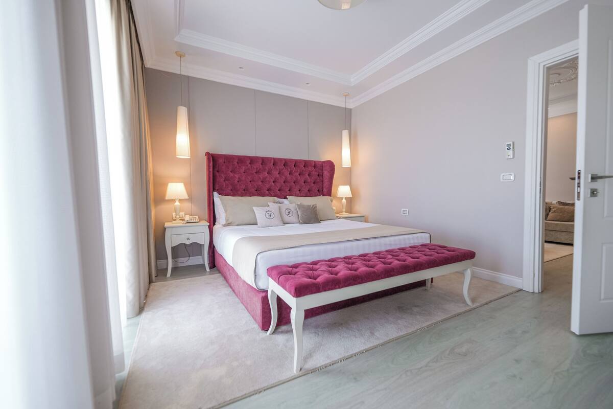 A bedroom with a large bed, bedside tables, a bench, and a door leading to another room.