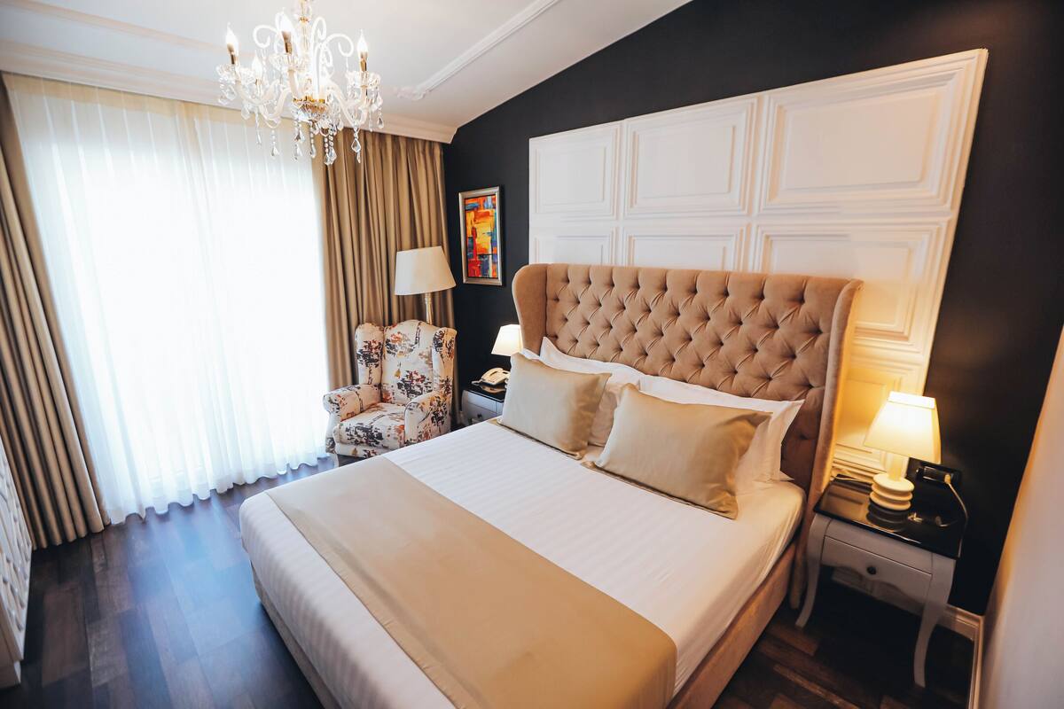 A hotel room with a large bed, a chandelier, a nightstand, a chair, and a painting on the wall.