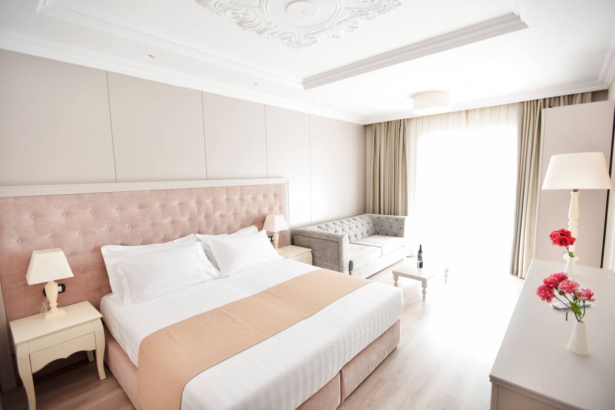 A hotel room with a large bed, a sofa, a nightstand with a lamp, and a vase with flowers.