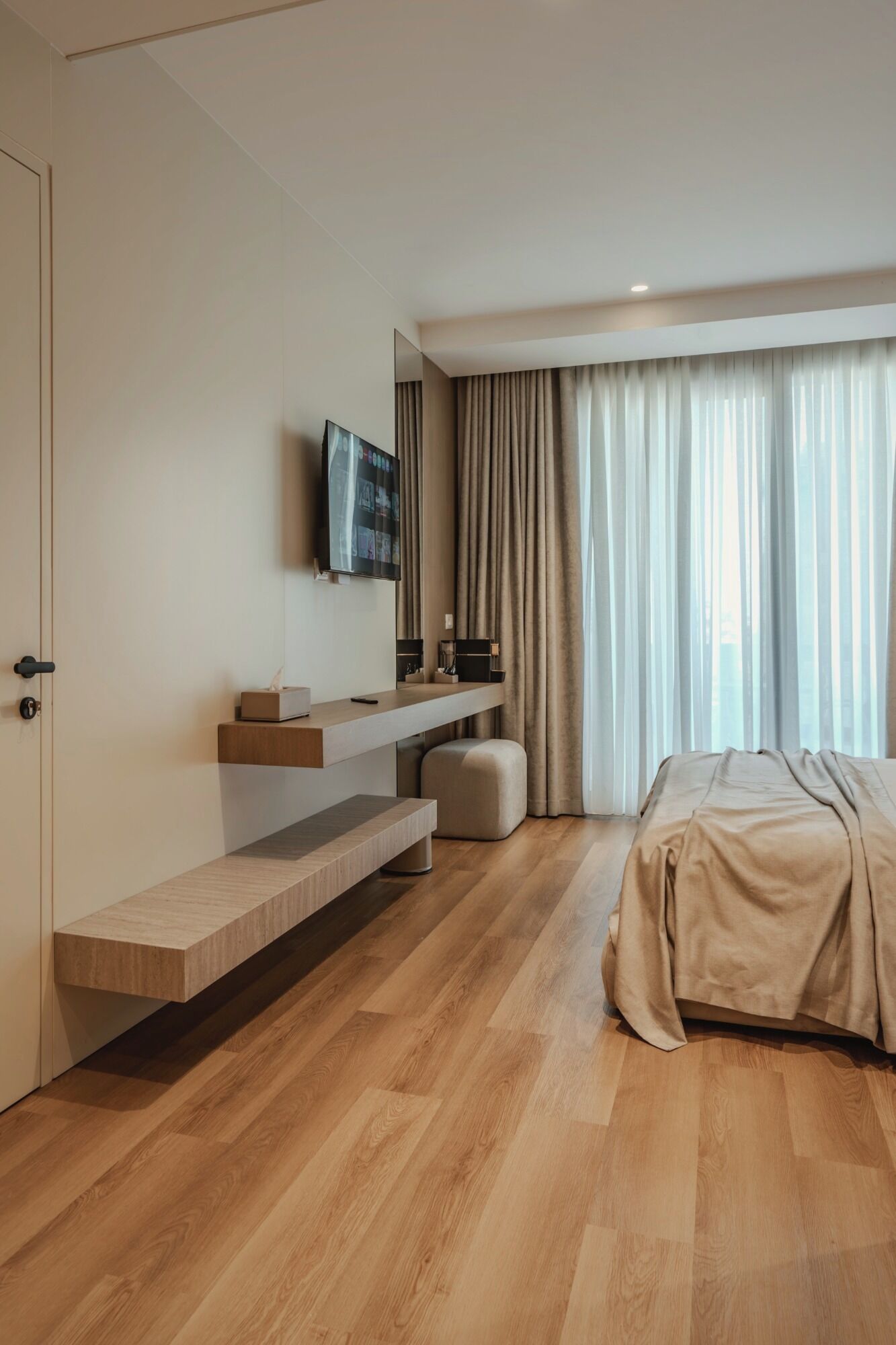 A modern hotel room with a wooden floor, a bed with a beige cover, a TV mounted on the wall, a bedside table with a remote, and a large window with curtains.