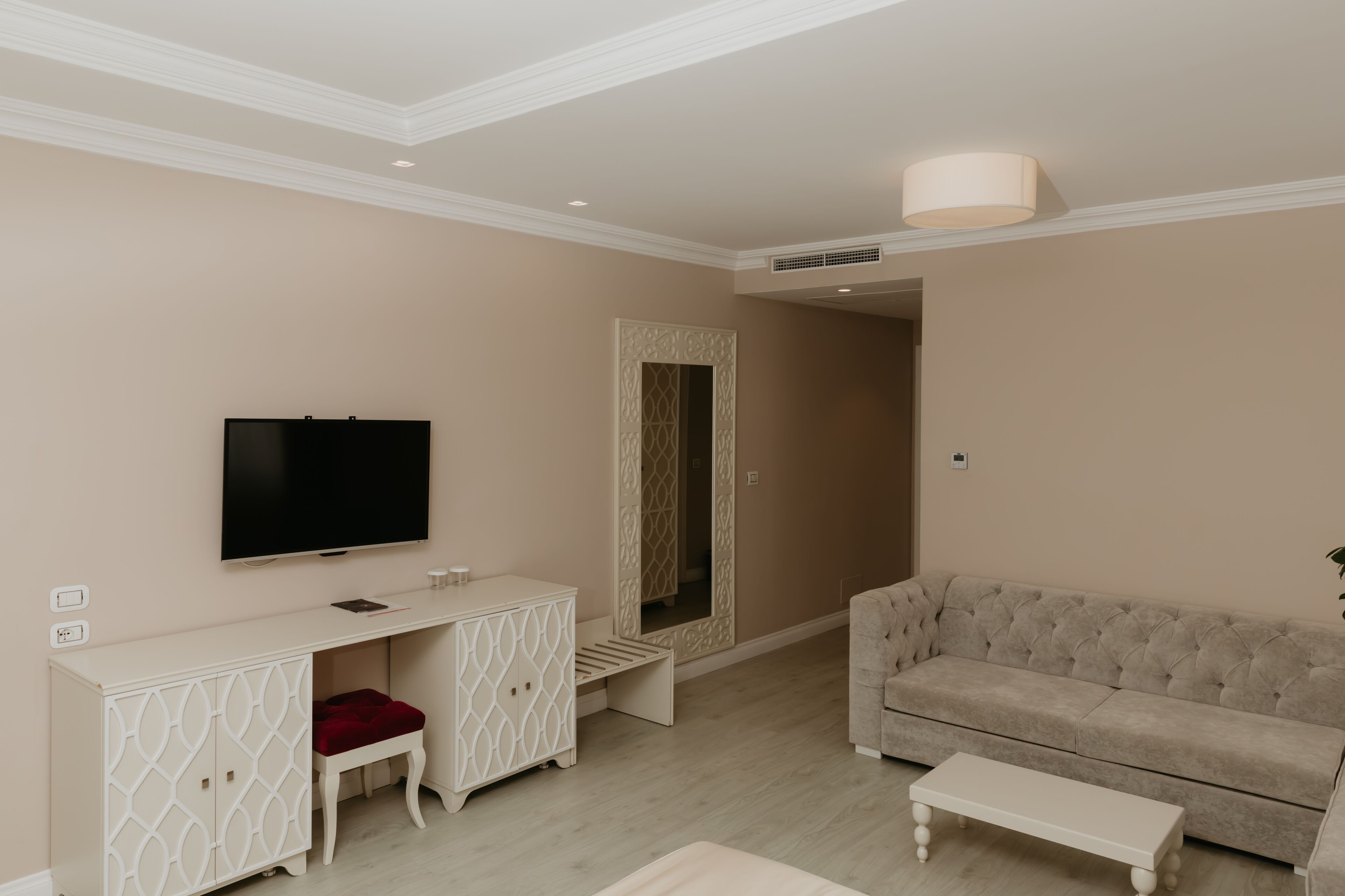 Un salon comprenant un téléviseur à écran plat, une console blanche, un tabouret rouge à coussin, un canapé beige et une table basse blanche.