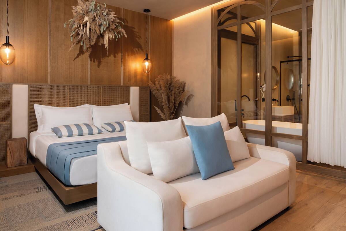 A modern bedroom with a large bed, a sofa, a glass shower area, and a wooden wall panel.