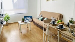 Ein gemütliches Wohnzimmer mit einer Couch, einem Couchtisch und einem Esstisch mit Stühlen. An den Wänden hängen zwei Gemälde und im Raum steht ein Blumentopf.