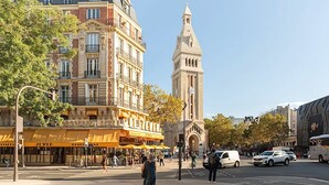 Un edificio storico con una torre dell'orologio, una strada con passanti e un ristorante con tende gialle.