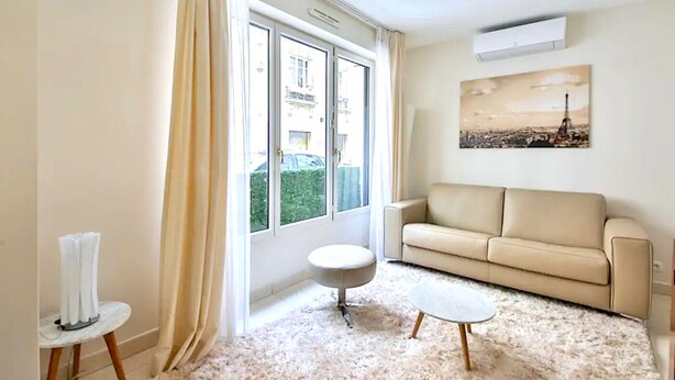 Un soggiorno con un divano color beige, un tavolino rotondo e un piccolo tavolino laterale. Si vede una finestra con tende, un quadro che raffigura uno skyline e un condizionatore d'aria a parete.