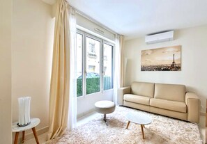 A living room with a beige sofa, a round coffee table, and a small side table. There is a window with curtains, a painting of a cityscape, and an air conditioning unit on the wall. - L'appartement Thibaud (Paris)
