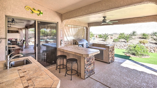 A covered outdoor kitchen area with a bar stool, a grill, and a view of a landscaped garden. - Heated Pool & Game Room: Lake Havasu Retreat (Lake Havasu City)