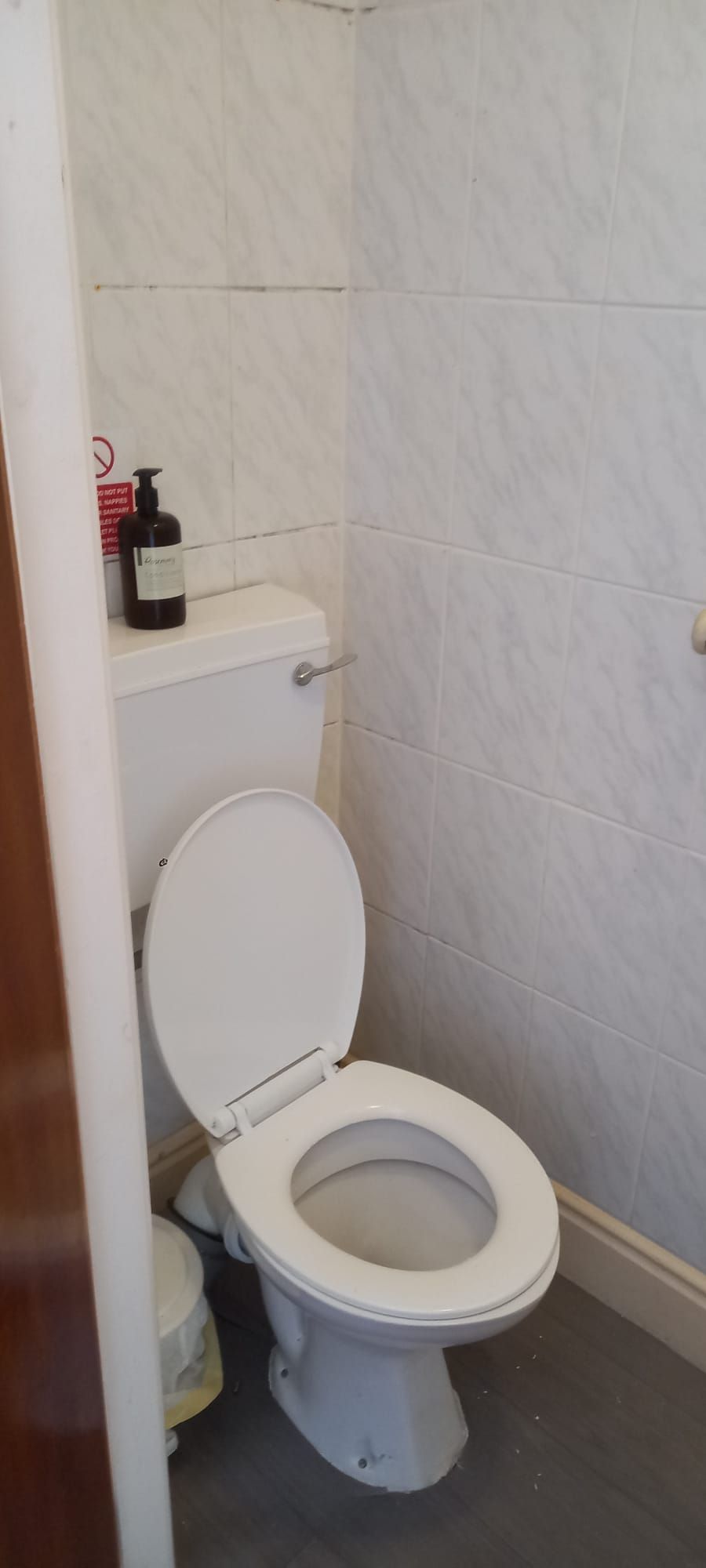 Ein Badezimmer mit einer weißen Toilette, einem Ablagefach mit einer Flasche und einer gefliesten Wand.
