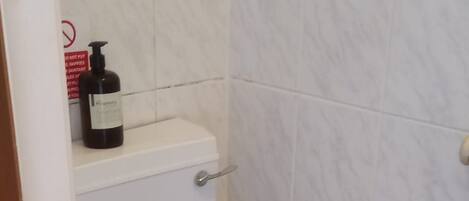 Ein Badezimmer mit einer weißen Toilette, einem Ablagefach mit einer Flasche und einer gefliesten Wand.