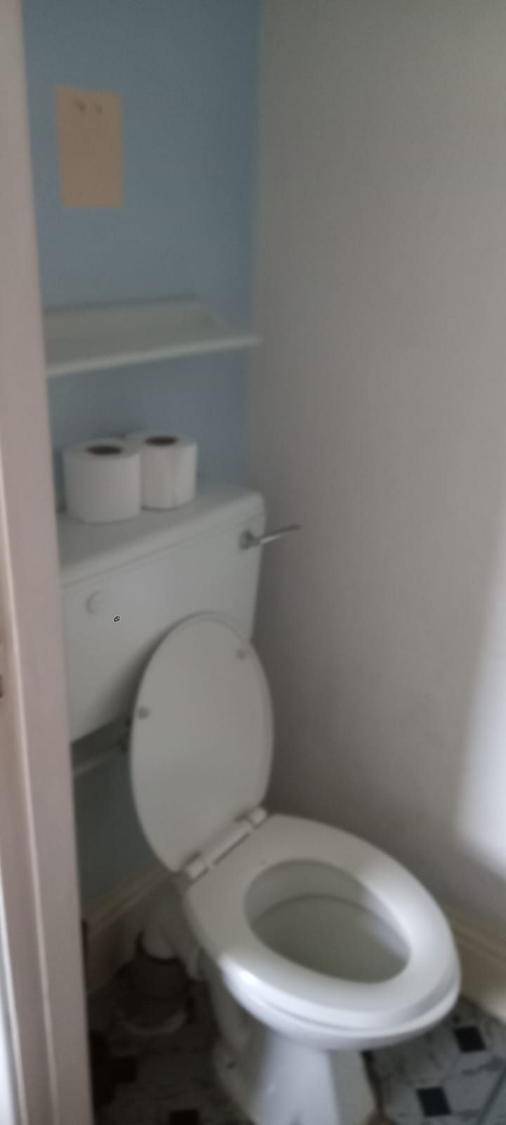 A small bathroom with a toilet, a shelf with toilet paper, and a wall with a small sign.