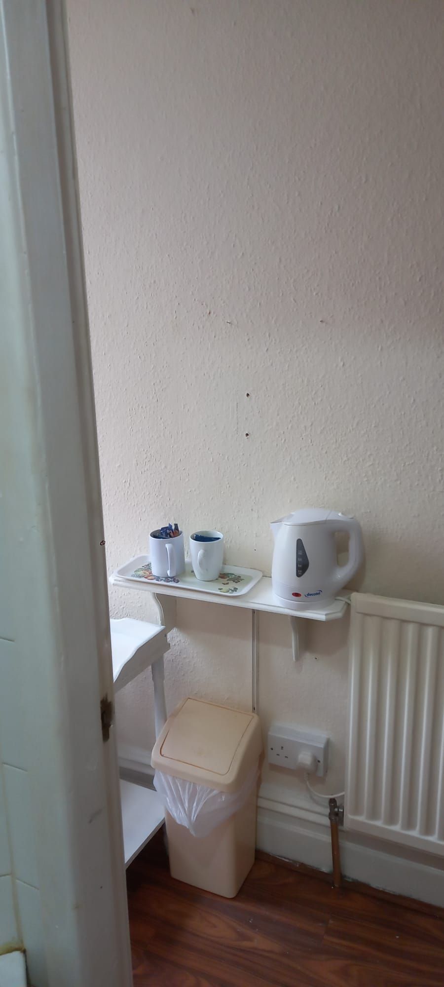 Una pequeña kitchenette con encimera blanca, hervidor y un cubo de basura.
