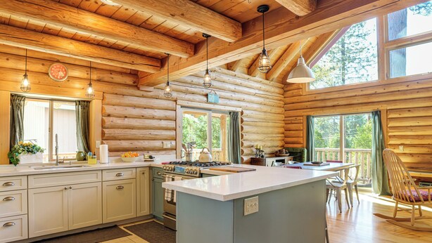 A log cabin interior with a kitchen featuring a central island, wooden beams, and large windows. - Walk to Pine Mountain Lake: Spacious Family Cabin (Groveland)