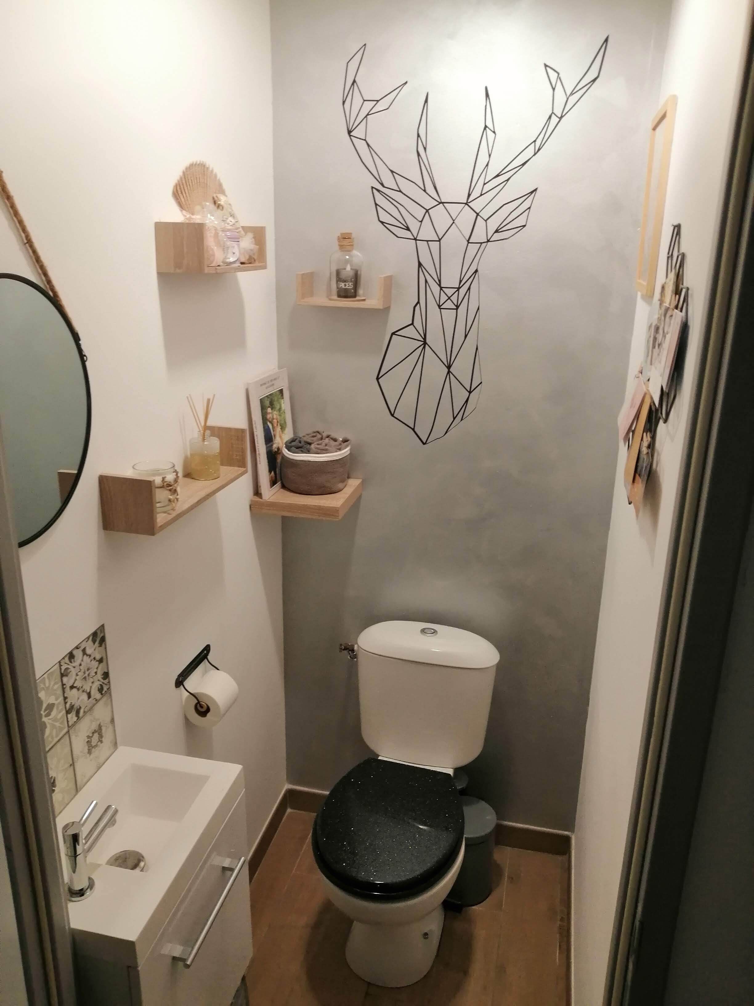 Baño pequeño con inodoro, lavamanos y espejo. Se observan repisas con artículos de aseo personal y un adhesivo en la pared.