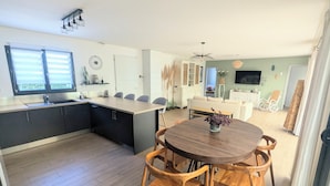Una cucina moderna con isola centrale, zona pranzo con sedie di legno e un soggiorno con divano e televisione.