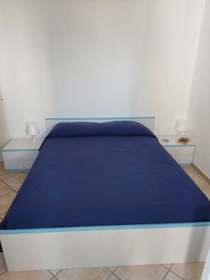 Un lit avec un matelas bleu et un cadre blanc, deux tables de chevet avec des lampes et une petite table de nuit avec un livre et une lampe.