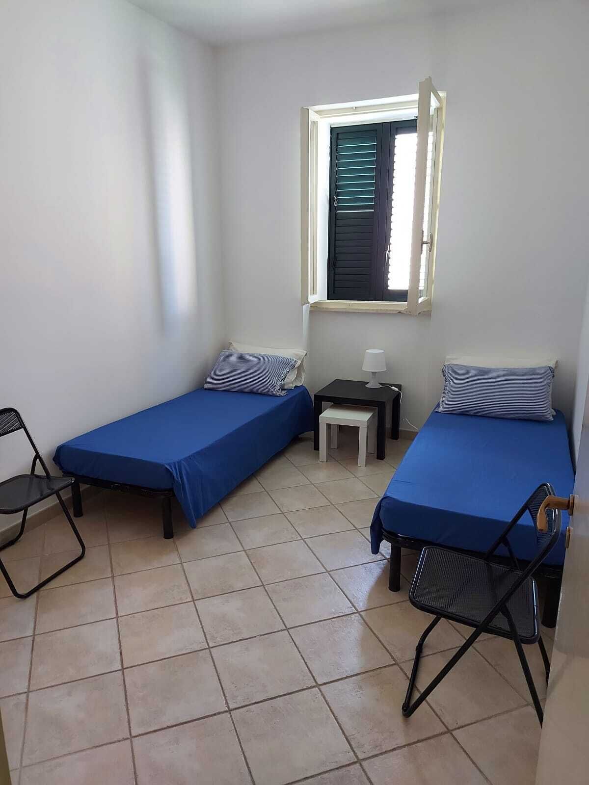 Une petite chambre avec deux lits simples, une fenêtre avec des stores, une petite table avec une lampe et une chaise.