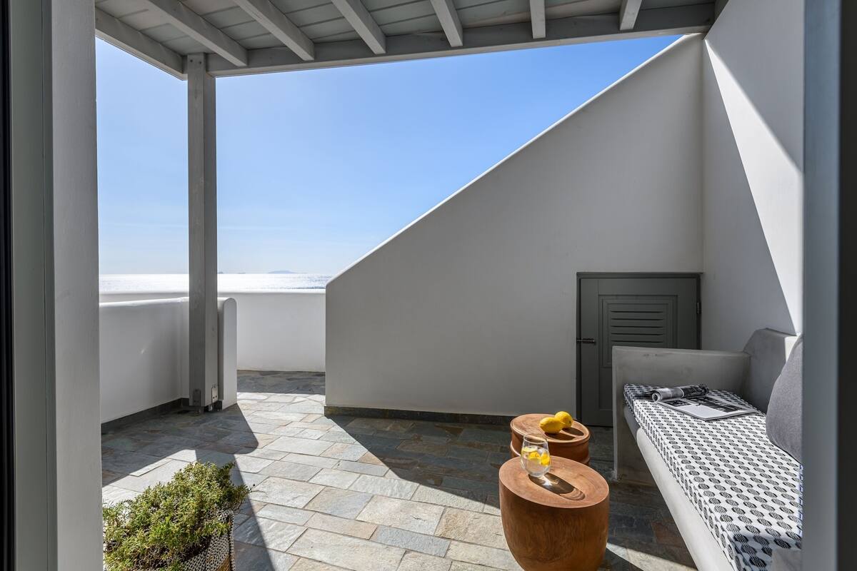 Una terraza en la azotea con un muro blanco, un banco de madera, una mesa pequeña con un vaso y limones, y vistas al mar.
