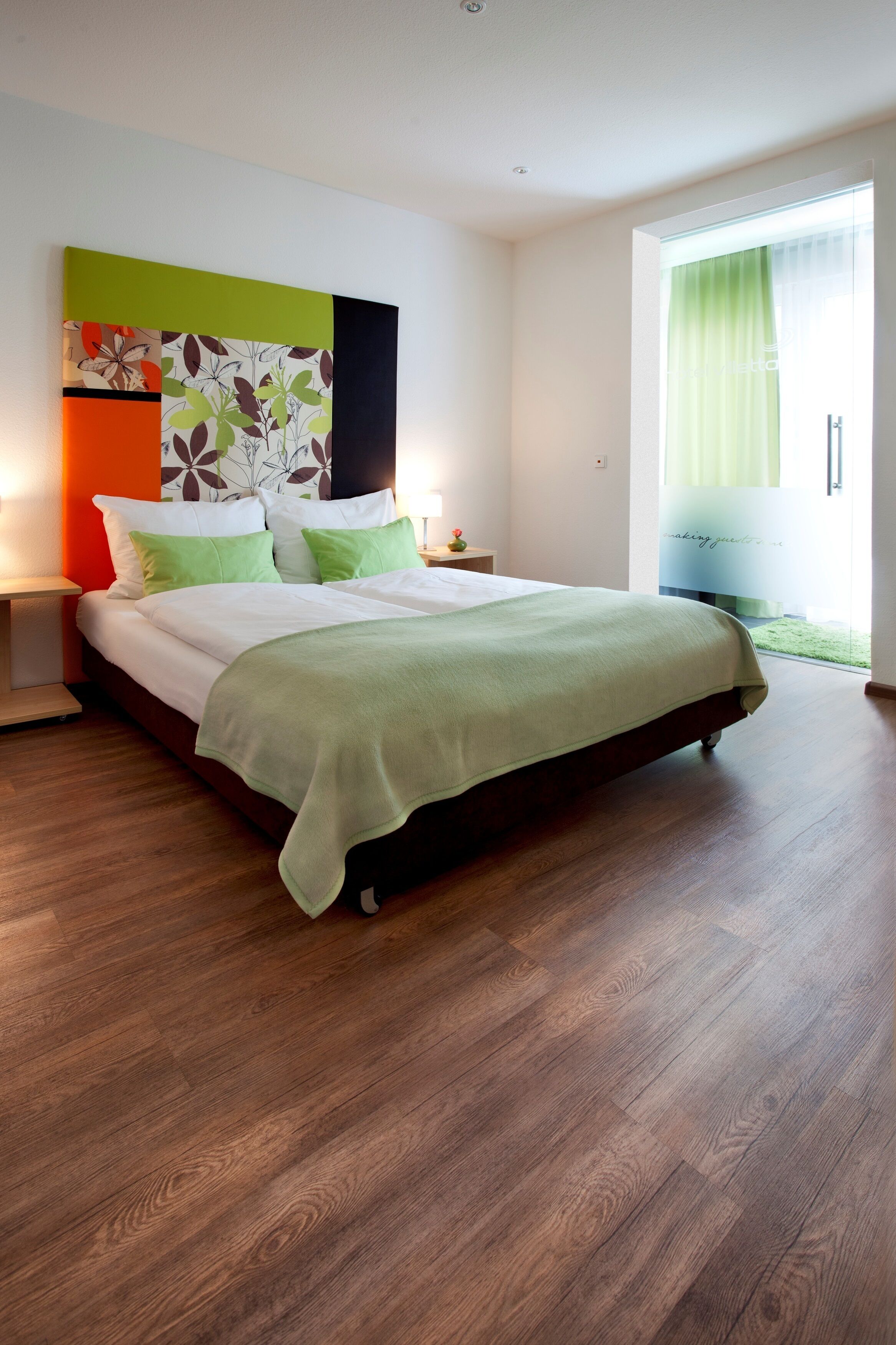 Un dormitorio moderno con una cama grande, ropa de cama verde, un adorno floral en la pared y vistas a un jardín verde a través de una ventana.