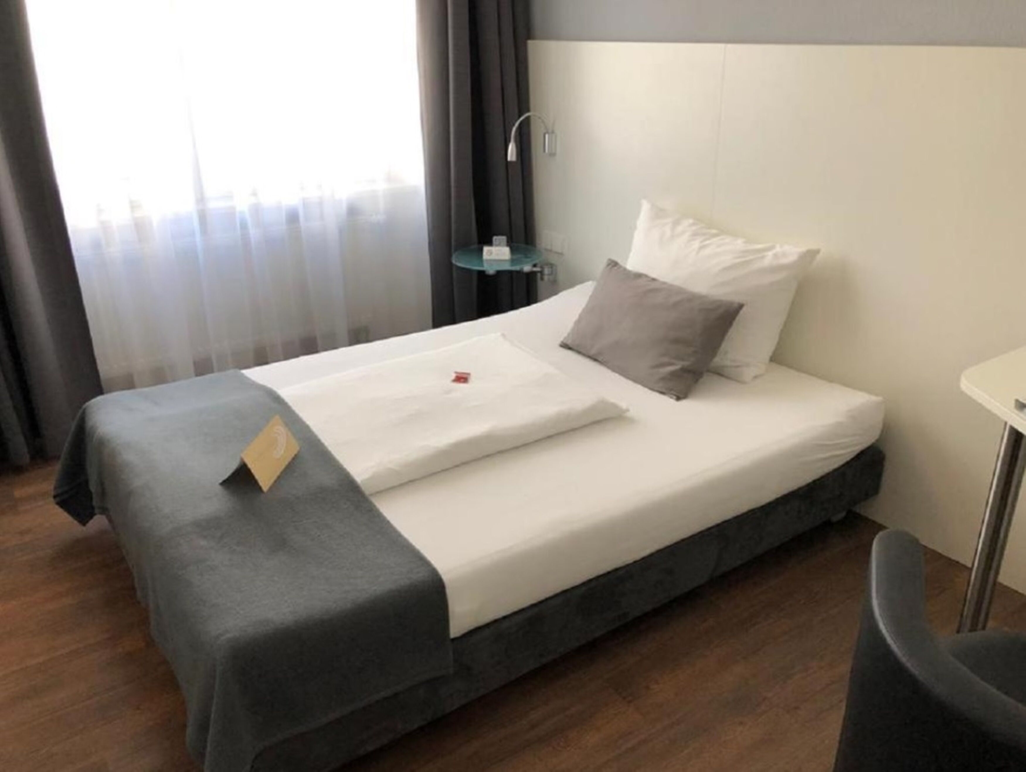 A single bed with a grey bedspread and white linens, a small bedside table with a lamp, and a view of the city through sheer curtains.
