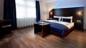 A modern hotel room with a wooden floor, a bed with white and dark blue bedding, a bedside table with a bottle and glass, and a nightstand with a lamp. - Schlotzeria Hotel I Cafe I Eis (Grenzach-Wyhlen)