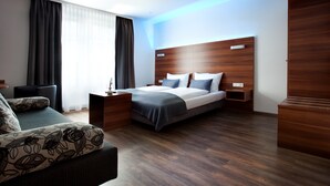 A modern hotel room with a wooden headboard, a bed with grey bedding, a dark sofa with floral cushions, and a wooden nightstand with a lamp. - Schlotzeria Hotel I Cafe I Eis (Grenzach-Wyhlen)