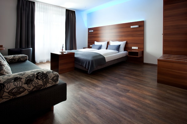Una habitación de hotel moderna con cabecera de madera, cama con ropa de cama gris, sofá oscuro con cojines estampados y mesita de noche de madera con lámpara.