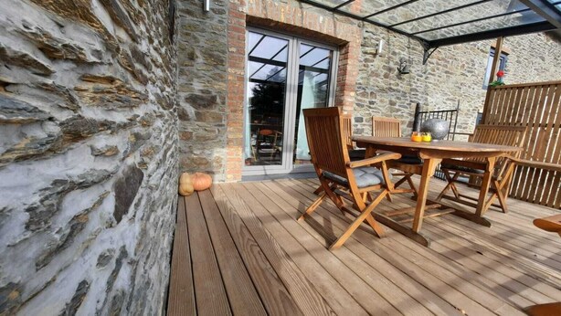 Eine Holzterrasse mit einer Steinmauer, einem Tisch mit Stühlen und einer Glastür zum Innenbereich.