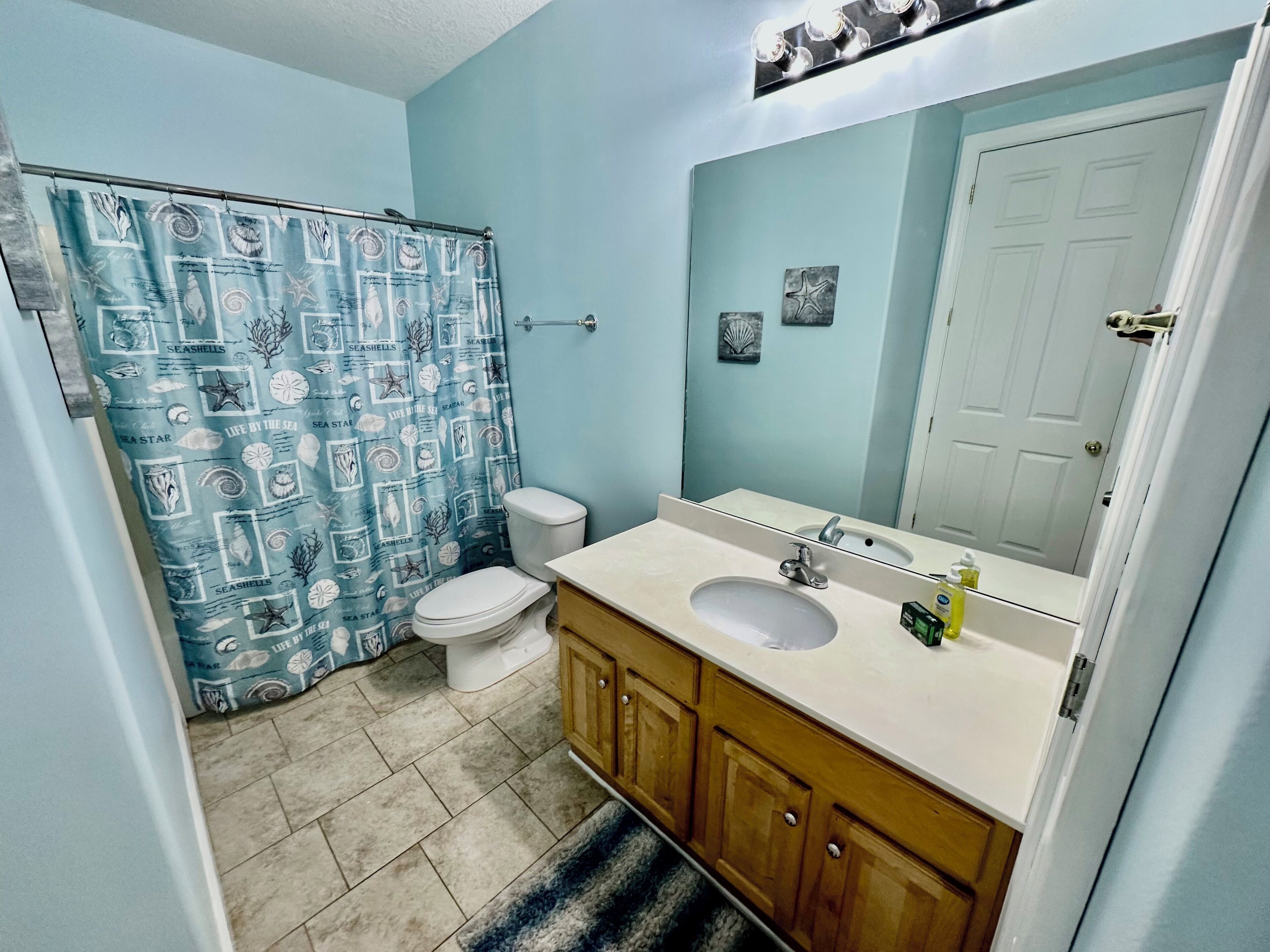 Ein Badezimmer mit einer blauen Duschvorhang mit Meeresmotiven, einer weißen Toilette, einem Waschbecken mit Spiegel, Holzschränken und einem gefliesten Boden.