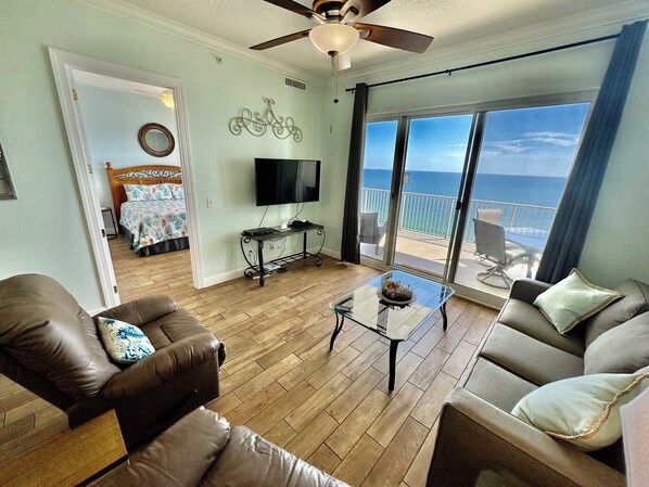 A spacious living room with a balcony, a ceiling fan, a television, and a bed with floral bedding. - Beach-Front Condo, Comfortable Recliners, XL Balcony, Free Beach Chairs! (Panama City Beach)
