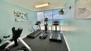 Sala de ginástica com esteiras, bicicleta ergométrica e remo, com vista para a praia através de amplas janelas.