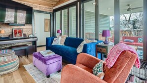 Una sala de estar moderna con techo de madera, ventanas grandes, un sofá azul, un sillón naranja y un reposapiés morado.