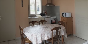 A dining area with a round table covered by a patterned tablecloth, surrounded by wooden chairs. A kitchen with a stove, microwave, and sink is visible in the background.