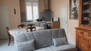 A cozy living room with a patterned sofa, a dining table with chairs, a kitchen area with a stove and microwave, and a wooden cabinet with glass doors.