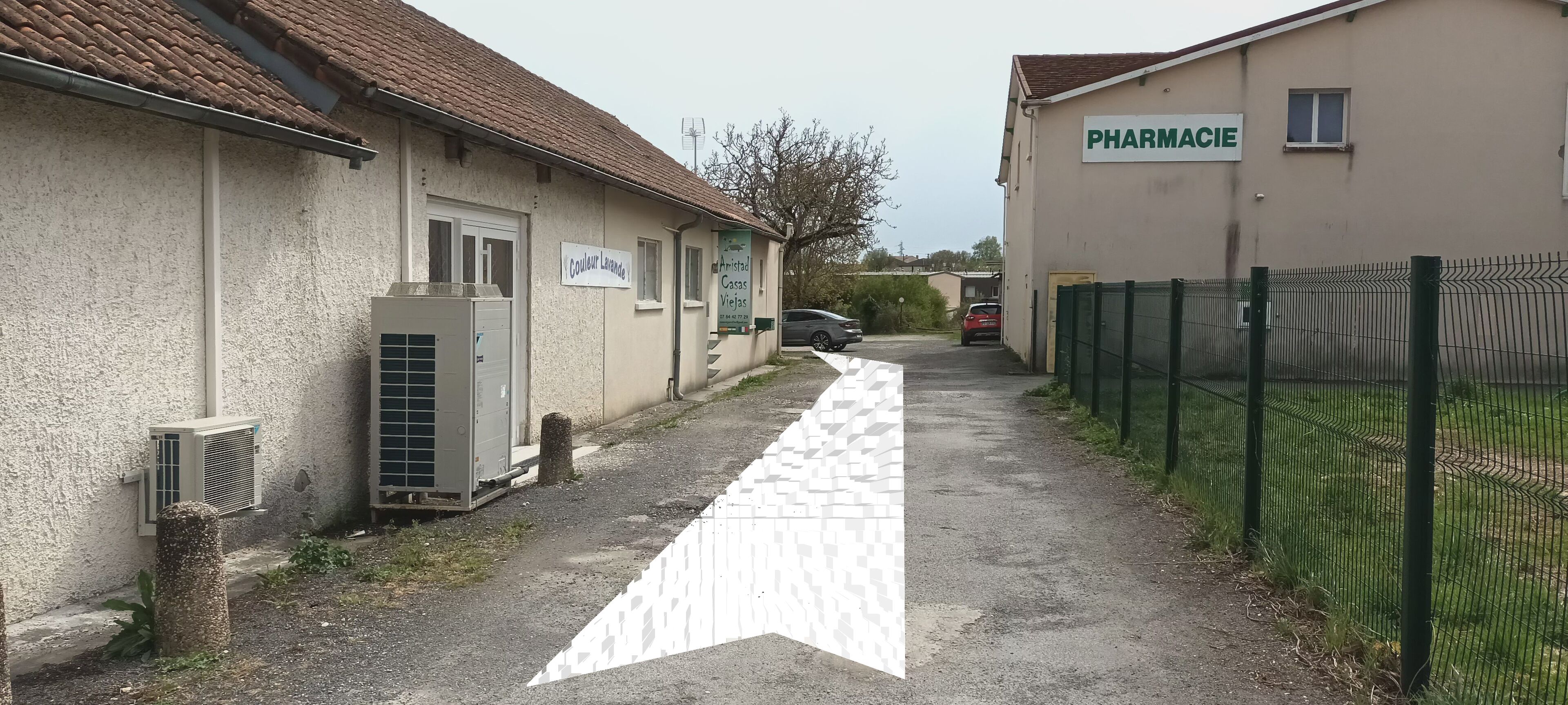 A narrow alleyway with a paved path, a building with a sign reading 'PHARMACIE', and a fenced area with parked cars.