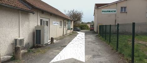 A narrow alleyway with a paved path, a building with a sign reading 'PHARMACIE', and a fenced area with parked cars.