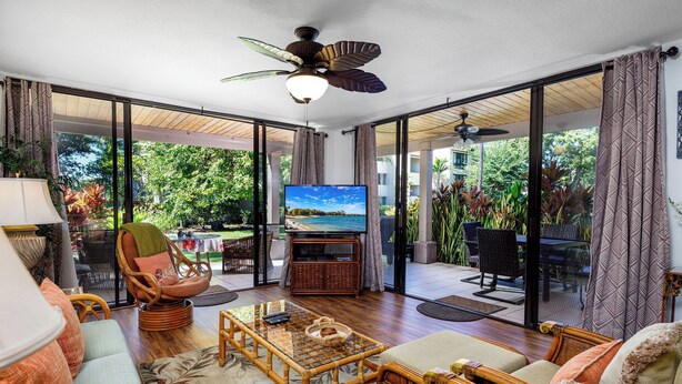 A spacious living room with a television, wicker furniture, and large sliding glass doors leading to an outdoor area. - KKSR#8101 GORGEOUS 2 BEDR, GROUND FLOOR CORNER, REMODELED, AIR CONDITIONING (Kailua Kona)