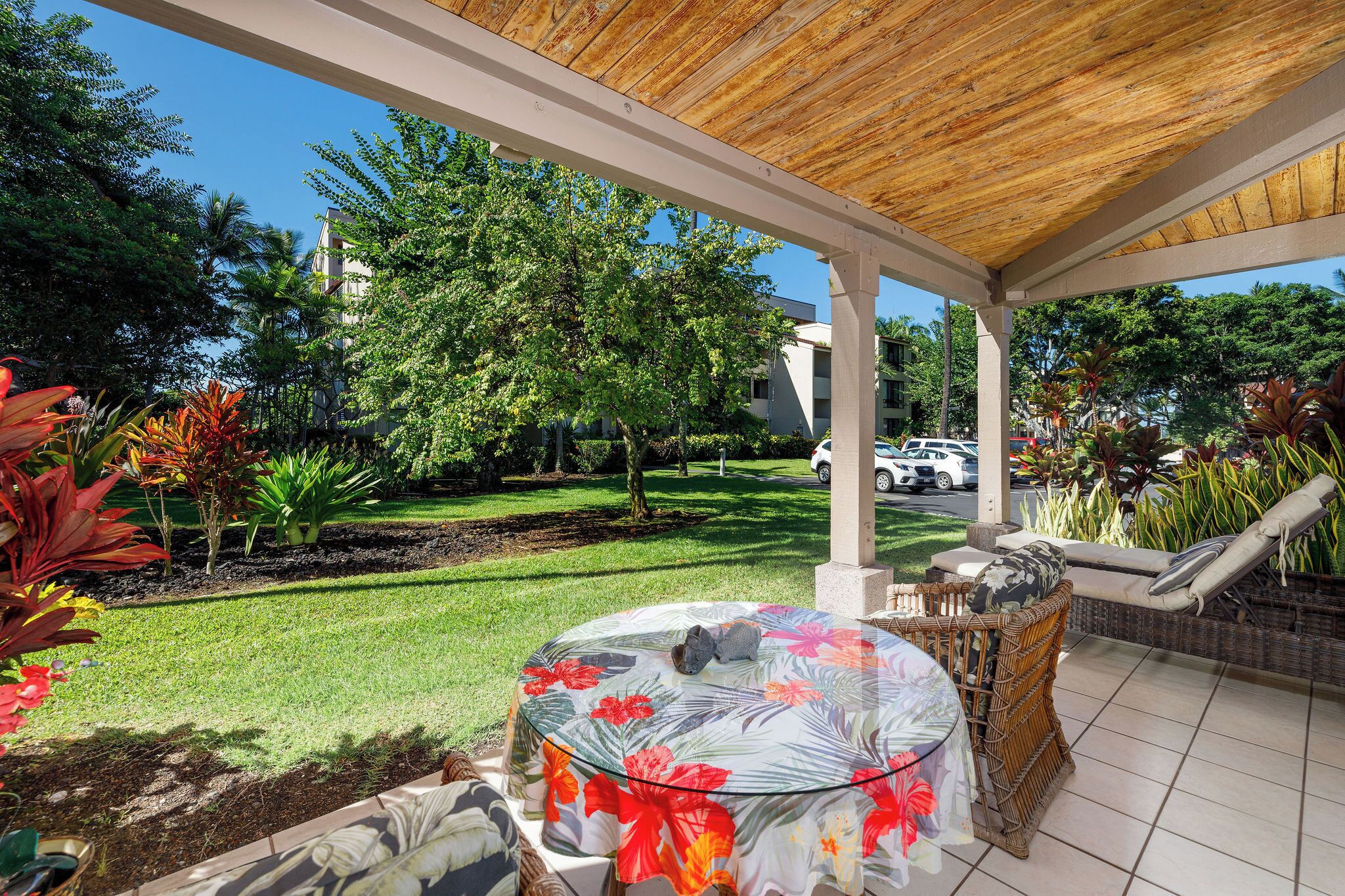 A covered patio with a round table and chairs, surrounded by lush greenery and a view of a parking area.
