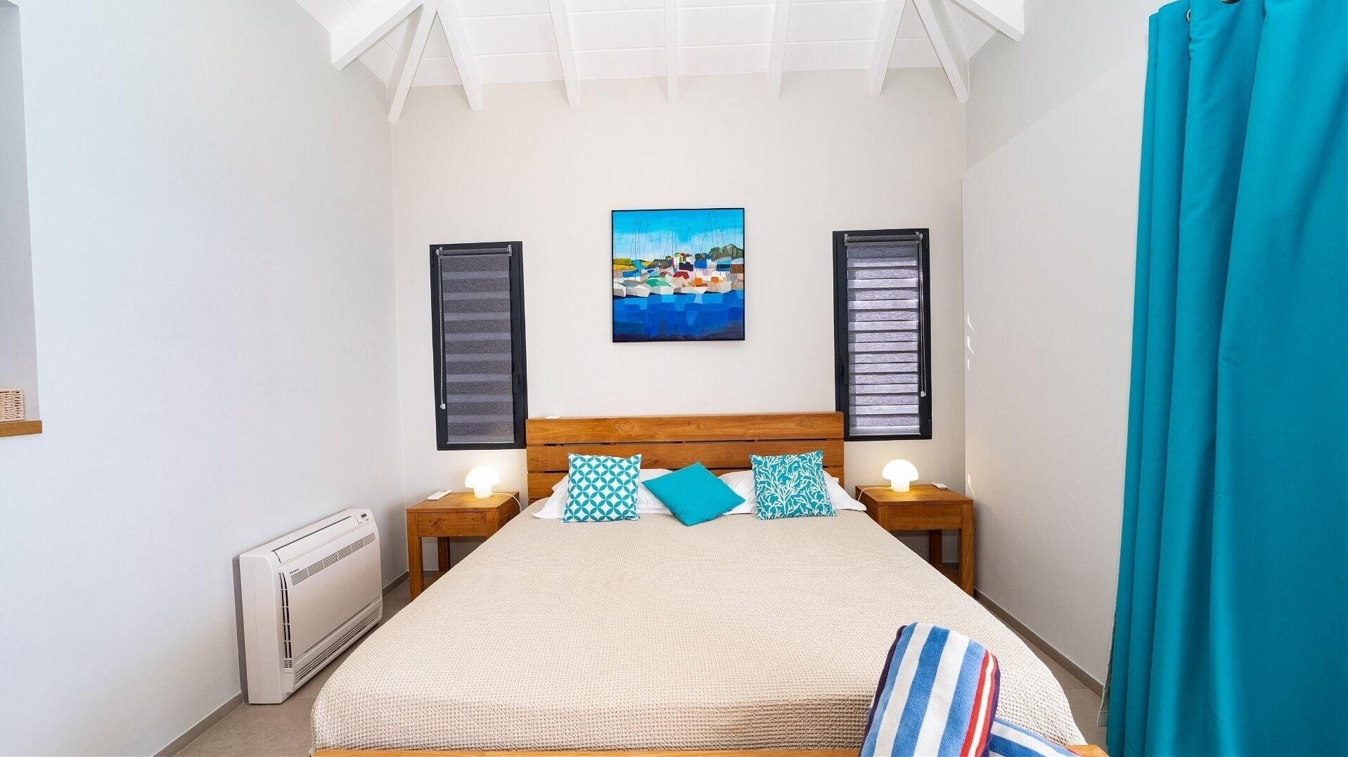 Un dormitorio con una cama, dos mesitas de noche con lámparas, un radiador, una ventana con contraventanas y un cuadro en la pared.