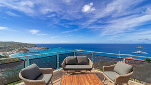 A balcony with wicker chairs and a wooden table overlooking a coastal view with boats and a distant island. - Belharra Luxury villa (Gustavia)