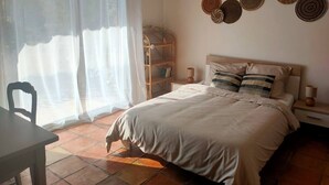 A bedroom with a bed, a bedside table, a chair, a window with sheer curtains, and a wall with woven wall art. - A bit of straw (Cagnes-sur-Mer)