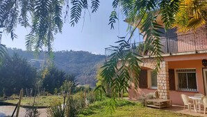 A two-story building with a balcony, surrounded by greenery and a view of a hilly landscape. - A bit of straw (Cagnes-sur-Mer)