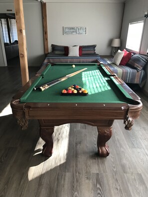 Ein Billardtisch mit Kugeln und Spielstöcken, eine Couch mit Kissen und ein Schild an der Wand mit der Aufschrift 'Welcome to LAKE'.