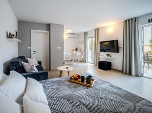 A modern living room with a sofa, a coffee table with a tray of fruit and mugs, a TV mounted on the wall, and a dining area with chairs and a table. - Apartments Tomy (Sutivan)