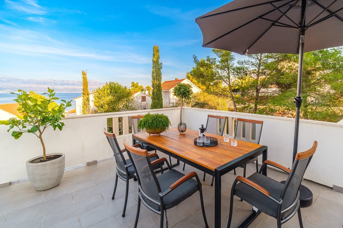 Una terraza con una mesa y sillas de madera, una planta en maceta y vistas al mar y edificios.