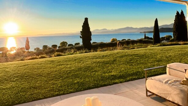 A circular outdoor seating area with a white table and a candle centerpiece, overlooking a grassy landscape and a body of water during sunset. - Stunning new villa with beautiful lake view (Bardolino)