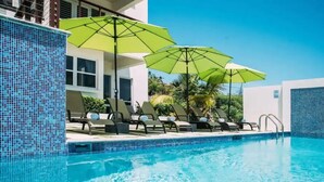 Ett poolområde med solstolar och parasoller, en pool med kakelbeklädd botten och en byggnad med stora fönster.