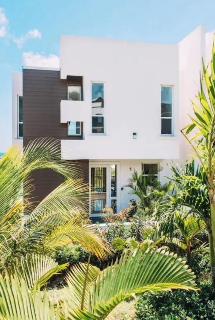 Une maison moderne à deux étages, dotée d’un toit plat, de grandes fenêtres et d’un jardin bien entretenu où poussent différentes espèces de plantes.