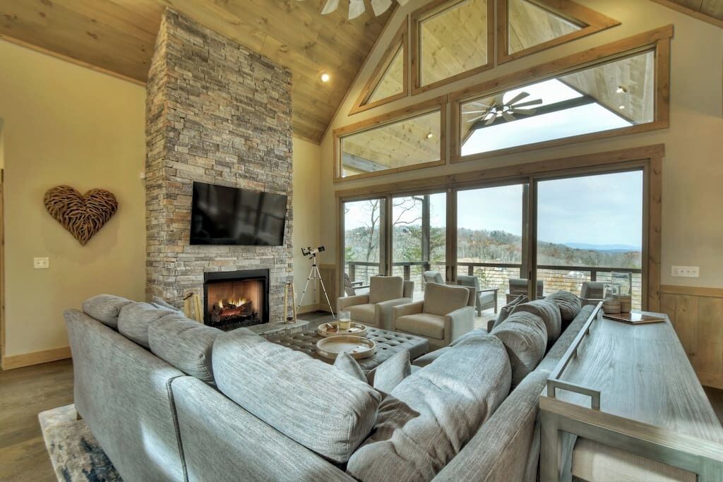 A spacious living room with a stone fireplace, comfortable seating, and large windows offering a view.
